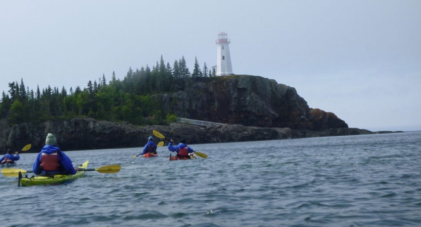a group of people paddling kayaks paddle toward a white lighthouse sitting atop a tall rocky shore